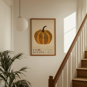 May include: A framed art print featuring a stylized orange pumpkin with black spots and a black stem. The print includes the text "YAYOI KUSAMA" and Japanese characters. The artwork is displayed on a white wall near a staircase.