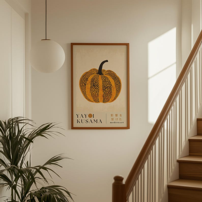 May include: A framed art print featuring a stylized orange pumpkin with black spots and a black stem. The print includes the text "YAYOI KUSAMA" and Japanese characters. The artwork is displayed on a white wall near a staircase.