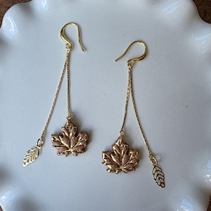 May include: Gold-toned dangle earrings featuring maple leaf and leaf charm pendants. The earrings have a hook closure and are displayed on a white, scalloped-edge plate. The background is a wooden surface.
