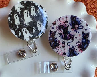 May include: Two Halloween-themed retractable badge reels. One features white ghosts on a black background, the other has black cats, skulls, and floral elements on a white background. Both have clear plastic clips.