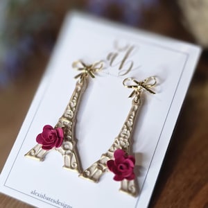 May include: Gold-colored Eiffel Tower earrings with a small red rose accent. The earrings are on a white card with the text "alexisbatesdesigns.etsy.com". Each earring has a small gold bow at the top.