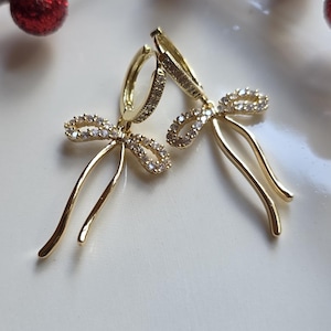 May include: A pair of gold-colored bow earrings with sparkling clear stones. The earrings are on a white surface, with red berries in the background. The earrings have a delicate, elegant design.