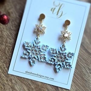 May include: A pair of snowflake earrings. Each earring features a light blue snowflake design with gold accents and a gold-colored stud. The earrings are displayed on a white card with the text alexisbatesdesigns.etsy.com.