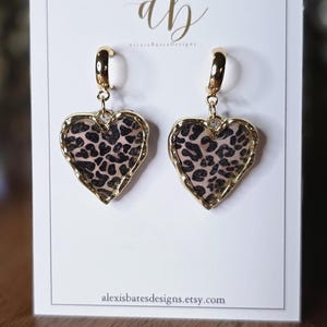 May include: A pair of heart-shaped earrings with a gold-colored trim and a leopard print design. The earrings are hanging on a white card with the Alexis Bates Designs logo and Etsy shop URL.