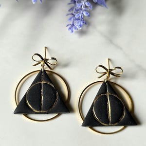 May include: A pair of black triangle earrings with a gold-colored Deathly Hallows symbol and a gold bow detail. The earrings are suspended from gold hoops. The background includes purple flowers and a white surface.