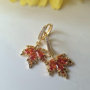 May include: A pair of gold-toned earrings featuring a maple leaf design. The leaves are adorned with orange and red gemstones. The earrings have a hoop closure and are set against a white background with a yellow flower.