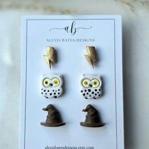 May include: A white card displays six pairs of earrings. The top row features gold lightning bolt studs. Below are white owl studs with black spots and yellow eyes. The bottom row shows brown witch hat studs. The text "alexisbatesdesigns.etsy.com" is at the bottom.