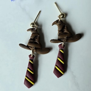 May include: A pair of brown polymer clay earrings. Each earring features a brown sorting hat charm and a burgundy tie charm with yellow stripes. The earrings have gold-colored ear wires.