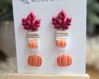May include: A white card displays handmade earrings with fall-themed designs. The earrings include burgundy maple leaves, coffee cups with pumpkins, and orange pumpkins. The card is on a small wooden easel, with the text "handmade ALEXIS BATES".