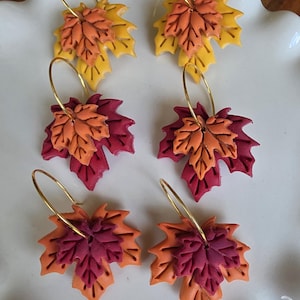 May include: Six pairs of fall-themed earrings. Each earring features a layered maple leaf design in shades of yellow, orange, and burgundy. The leaves are attached to gold-colored hoop earrings. The earrings are displayed on a white, scalloped plate.