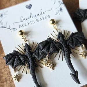 May include: Black dragon earrings with gold accents and star charms. The earrings are handmade and feature a detailed dragon design with outstretched wings. The earrings hang from gold-colored ball studs.
