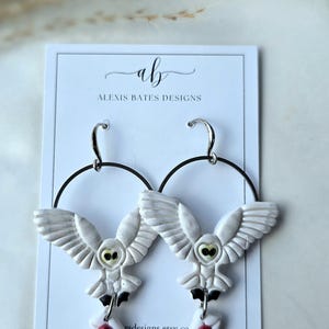 May include: White owl earrings with black hoop accents. Each earring features a detailed owl design with outstretched wings, a small envelope, and a red heart. The earrings are displayed on a white card with the Alexis Bates Designs logo.