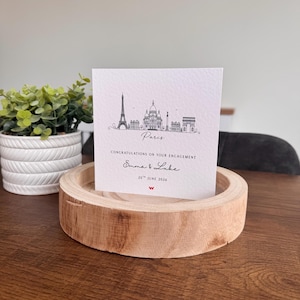 Puede incluir: Una tarjeta de felicitación cuadrada blanca con un dibujo de línea negra del horizonte de París, incluida la Torre Eiffel. El texto dice: Paris, CONGRATULATIONS ON YOUR ENGAGEMENT, Emma & Luke, 25th JUNE 2026, y un pequeño logotipo rojo abajo.