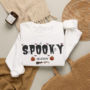 Puede incluir: Una sudadera blanca de manga larga con el texto negro SPOOKY SEASON. El diseño incluye pequeñas ilustraciones de fantasmas, murciélagos, calabazas naranjas y una escoba de bruja dispersas alrededor de las letras en la parte delantera.