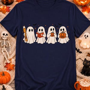 Puede incluir: Una camiseta azul marino de manga corta con un estampado gráfico de cuatro fantasmas blancos. Los fantasmas sostienen una escoba, un libro naranja, una taza naranja humeante y una calabaza tallada, rodeados de pequeñas estrellas y murciélagos naranjas.