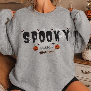 Puede incluir: Una sudadera gris jaspeado de cuello redondo con el texto negro SPOOKY en una fuente que gotea, con la palabra season en letra cursiva debajo. El diseño incluye pequeñas calabazas naranjas, fantasmas blancos, murciélagos negros y una escoba.