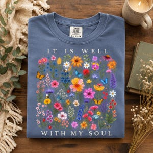 Puede incluir: Una camiseta azul de cuello redondo con un estampado botánico colorido de flores silvestres, mariposas y mariquitas. El texto "IT IS WELL WITH MY SOUL" está impreso en blanco sobre y debajo del arreglo floral en la parte frontal de la prenda.