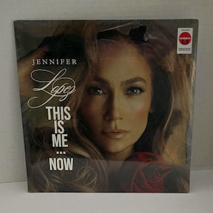 May include: A vinyl record album featuring Jennifer Lopez. The album cover has a close-up image of Jennifer Lopez's face with the text "Jennifer Lopez" and "This Is Me...Now" in white lettering. A red circle with the word "exclusive" is in the upper right corner.