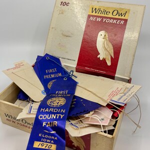 May include: A vintage White Owl New Yorker cigar box containing several blue First Premium ribbons from the 1970 Hardin County Fair in Eldora, Iowa. The box lid features an owl illustration and the text 10 cents, while the ribbons include strings.