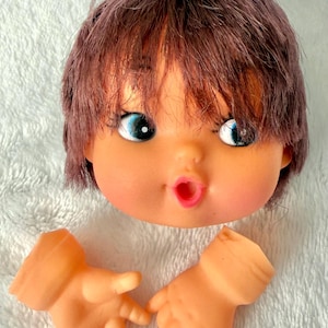 May include: A vintage doll head with brown hair, blue eyes, and an open mouth. Two detached doll hands are also visible. The doll's face is peach-colored, and the hair is styled in a layered cut.