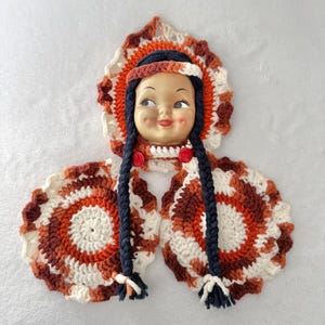 Puede incluir: Una cabeza de muñeca vintage con un tocado y cuello de ganchillo. El tocado presenta círculos concéntricos en tonos de naranja, marrón y crema. La muñeca tiene trenzas azul oscuro y detalles de botones rojos. El diseño general recuerda al arte nativo americano.