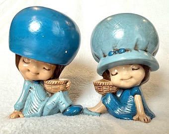 Könnte beinhalten: Zwei Keramikfiguren von Kindern in blauen Outfits und Hüten, die jeweils einen kleinen Korb halten. Die Figuren haben geschlossene Augen und befinden sich in einer entspannten Sitzposition. Die Farbpalette ist hauptsächlich blau und beige.