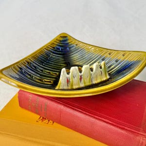 May include: A vintage, triangular ashtray with a yellow rim and a textured, glazed surface. The ashtray features a design of blue and yellow swirls and has four cigarette rests. It is resting on top of a red book.
