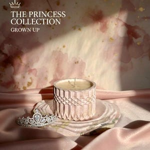 May include: A pink candle in a geometric patterned holder sits on a matching plate, next to a silver tiara. The candle is part of "THE PRINCESS COLLECTION" and is made of eco-resin pottery and soy wax.