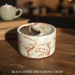 May include: A soy wax candle with a yin-yang design, featuring black coffee and almond crème scents. The candle is in a marbled white and brown container. The text "FIND YOUR BALANCE" is below the candle.