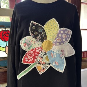 May include: A black long-sleeve sweatshirt featuring a large, multi-colored floral applique on the front. The flower is constructed from various patterned fabric patches with frayed edges, a solid yellow center, and a green fabric stem.