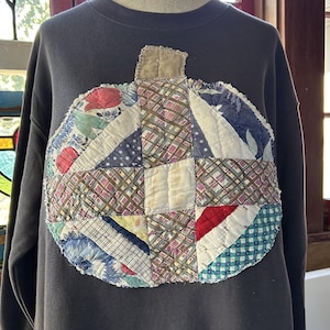 May include: A dark gray long-sleeve sweatshirt featuring a large, circular, pumpkin-shaped appliqué on the chest. The appliqué is constructed from various quilted fabric patches in assorted patterns and colors, including plaid, floral, and gingham, with a small tan fabric stem at the top.