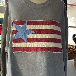 May include: A heather gray crewneck sweatshirt featuring a rectangular quilted patch on the chest. The patch displays a red and white striped pattern with a single light blue star on the left side, finished with raw, frayed edges.