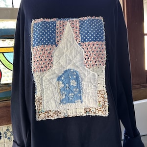 May include: A navy blue long-sleeve sweatshirt featuring a large, rectangular, quilted fabric appliqué on the front. The appliqué depicts a white church structure with a blue floral door, surrounded by patches of blue polka dot and floral fabric.