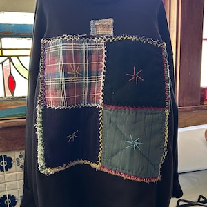 May include: A black long-sleeve sweatshirt featuring a large, square patchwork panel on the front. The panel consists of four quilted squares in plaid, solid black, and dark green, all joined with visible, decorative multi-colored stitching.