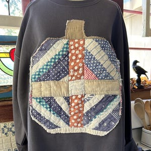 May include: A charcoal gray crewneck sweatshirt featuring a large, central pumpkin-shaped appliqué made from various vintage-style quilted fabric scraps in shades of blue, white, and orange with a frayed, raw-edge finish and a tan fabric stem.
