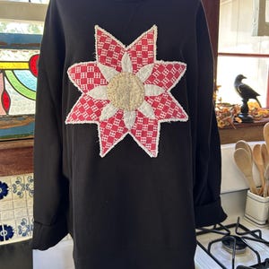 May include: A black long-sleeve sweatshirt featuring a large, centered star-shaped appliqué. The star is constructed from red and white checkered fabric with a textured, light-colored circular center and frayed white edges on the star points.