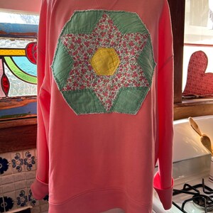 May include: A coral-colored sweatshirt with a crew neck and long sleeves. The front features a large, appliqued flower design. The flower has a yellow center, pink floral print petals, and green fabric surrounding the petals. The cuffs and hem are ribbed.