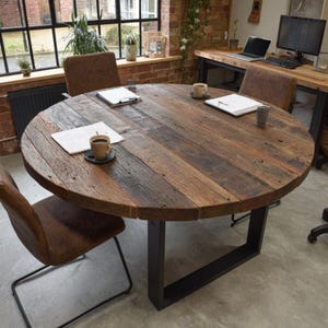Op de afbeelding: Een ronde houten tafel met een donkerbruine afwerking, ondersteund door een zwart metalen onderstel. Bruine leren stoelen omringen de tafel. Koffiekopjes en papieren liggen erop, wat wijst op een werkruimte.