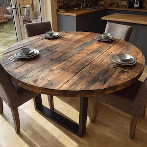 Op de afbeelding: Een ronde eettafel van gerecycled hout met een rustieke uitstraling. De tafel heeft een zwart metalen onderstel en is gedekt met vier couverts. De diameter van de tafel is ongeveer 122 cm.