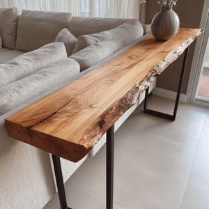 May include: A natural wood console table with a live edge and dark metal legs. The table is approximately 72 inches long and features a light brown finish. A decorative vase sits on the table.
