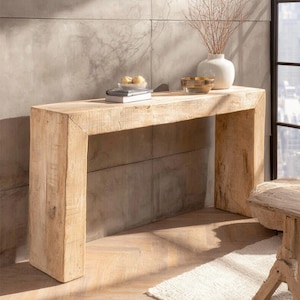 May include: A light-toned wooden console table with a natural finish. The table has a rectangular top and thick, blocky legs. A white vase with branches, a bowl, and books are displayed on the table's surface. The table is made of wood.