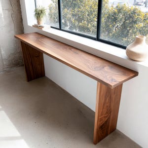 Puede incluir: Una consola de madera rectangular y larga con un acabado natural de vetas. La mesa está sostenida por dos patas gruesas y se coloca contra una pared y una ventana blancas. Una pequeña planta y un jarrón son visibles en el alféizar.