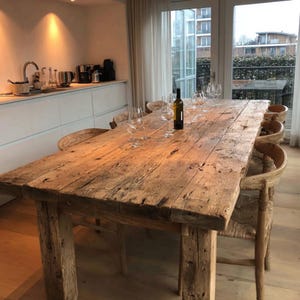 Può includere: Un grande tavolo da pranzo in legno rustico, lungo circa 240 cm, dall'aspetto vissuto. Il tavolo è circondato da sedie in legno con sedute intrecciate. Una bottiglia di vino e bicchieri sono sul tavolo. La stanza è luminosa.