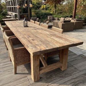 May include: A large, rectangular wooden outdoor dining table with a weathered, natural finish. The table is supported by thick, sturdy legs and is surrounded by woven wicker chairs. A small black lantern sits on the table's surface. The table measures approximately 305 cm long.