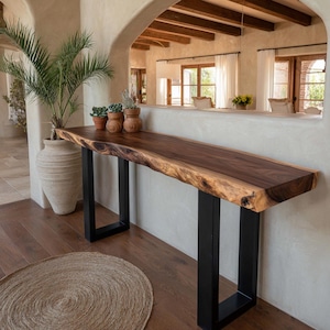Puede incluir: Consola de madera con borde natural, acabado marrón oscuro y patas de metal negro. La mesa mide aproximadamente 183 cm de largo y está colocada contra una pared neutra. Pequeñas plantas en macetas adornan la superficie.