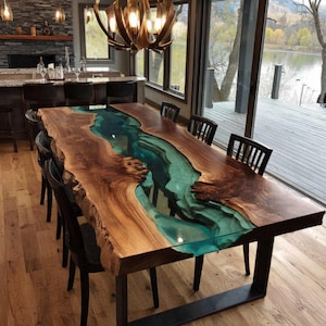 May include: A large dining table with a live edge design. The table has a teal-coloured resin river running through the centre of the walnut wood. It is supported by black metal legs and surrounded by black chairs. The table is approximately 240 cm long.