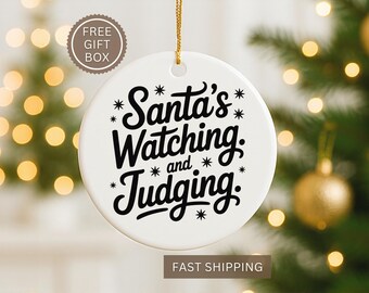 May include: A white ceramic Christmas ornament with the text "Santa's Watching and Judging" in black script. The ornament hangs from a gold string, with a blurred Christmas tree and lights in the background.