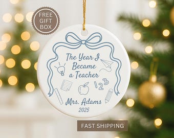May include: White ceramic ornament with a blue bow and the text "The Year I Became a Teacher." Includes teacher-themed doodles and "Mrs. Adams 2025." A gold string is attached for hanging. The ornament is a gift.