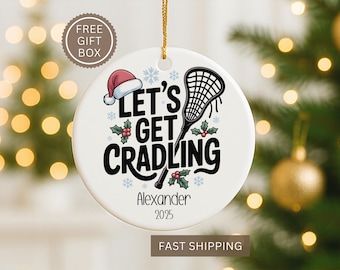 May include: White ceramic Christmas ornament with a lacrosse theme. The ornament features the text "Let's Get Cradling" with a Santa hat, snowflakes, and holly. Includes a lacrosse stick graphic and the name "Alexander" and the year "2025".