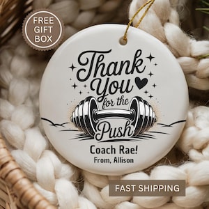 May include: A white ceramic ornament with a gold string for hanging. The ornament features the text "Thank You for the Push" with a barbell illustration. Below the text are the words "Coach Rae! From, Allison."